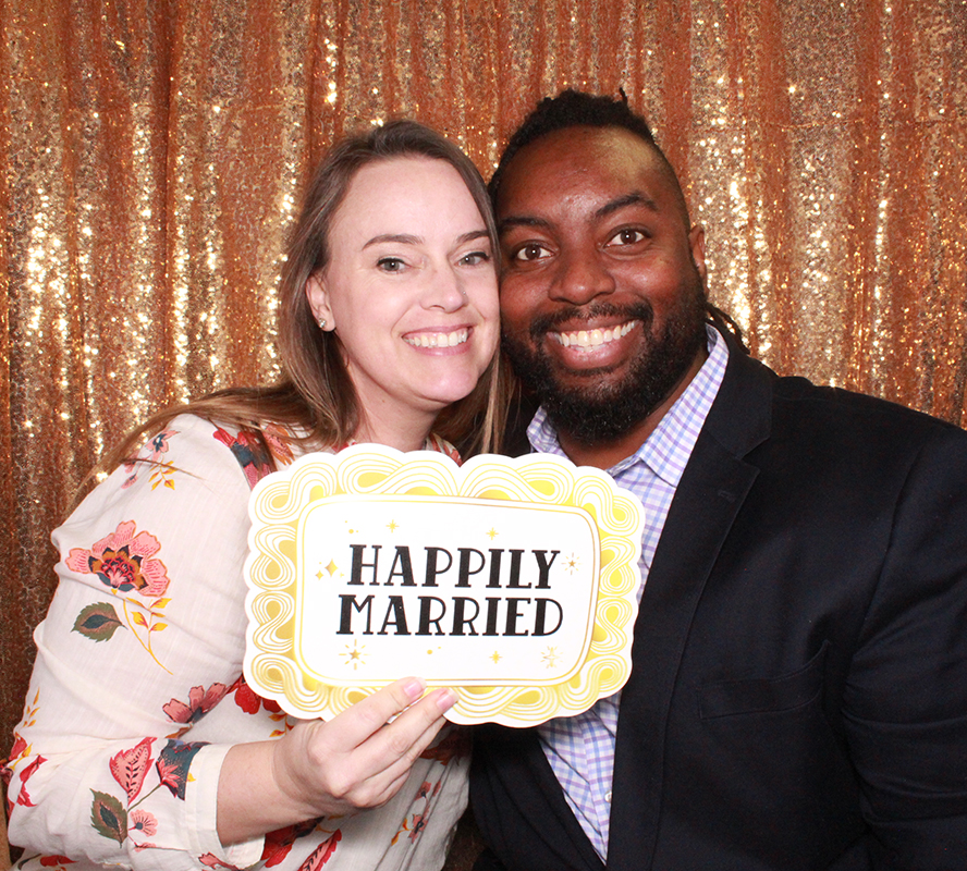 A cute couple holding the happily married photo booth prop with the gold sequin backdrop