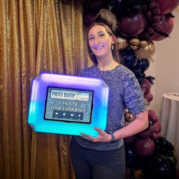 Woman holding a flashing mobile social media photo booth in front of a sparkly gold backdrop and balloon arch 