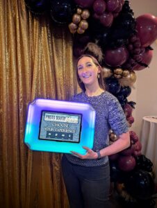 Woman holding a flashing mobile social media photo booth in front of a sparkly gold backdrop and balloon arch